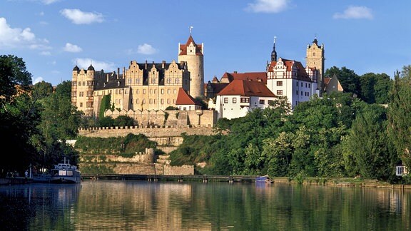 Blick über einen Fluss auf Schloss Bernburg, das über dem anderen Ufer thront, darüber blauer Himmel