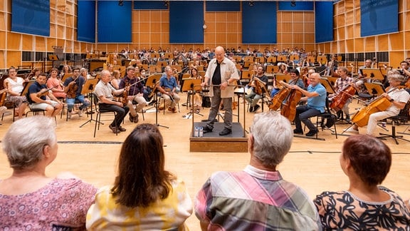 Vier Personen sitzen vor einem Orchester und Chor und sehen bei der Probe zu.