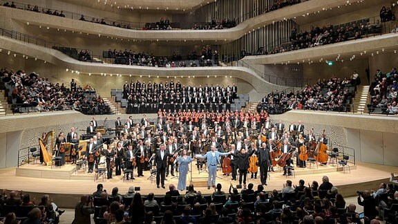 Alle drei MDR-Ensembles auf der Bühne der Hamburger Elbphilharmonie beim Applaus, davor fassen sich in vorderster Reihe Dirigenten und Solisten bei den Händen