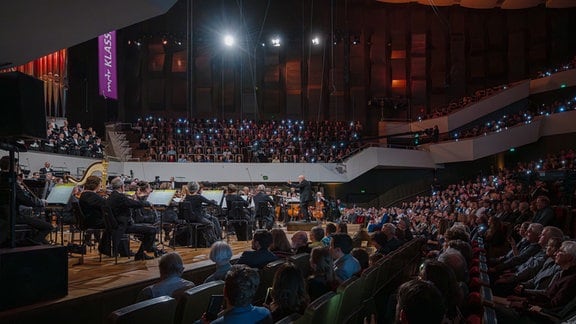 MDR-Sinfonieorchester und MDR-Rundfunkchor auf der Bühne des Leipziger Gewandhauses beim Festkonzert zum 100. Geburtstag der MDR-Ensembles, im Publikum leuchten Handy-Lichter.