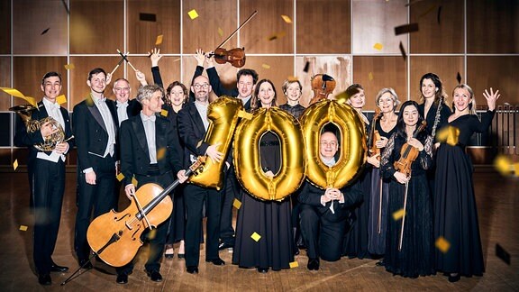 Mitglieder von MDR-Sinfonieorchester und MDR-Rundfunkchor in Konzertkleidung mit Instrumenten, Konfetti und einer goldenen 100 aus Folienballons.