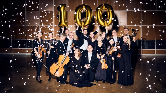 Mitglieder von MDR-Sinfonieorchester und MDR-Rundfunkchor in Konzertkleidung mit Instrumenten, Konfetti und einer goldenen 100 aus Folienballons.
