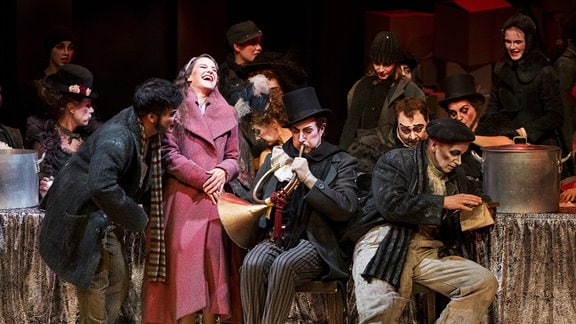 Szenenbild mit vielen Menschen auf einer Bühne, die an Tische gelehnt stehen oder sitzen und lachen, die meisten von ihnen Männer in dunklen Anzügen, in der Mitte eine Frau in rotem Mantel