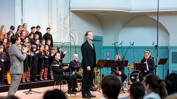 Auf einer Bühne stehen der MDR-Kinderchor, ein Instrumentalensemble, Dirigent Alexander Schmitt und Moderator Juri Tetzlaff.