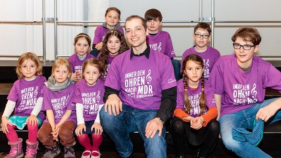 Kinder des MDR-Kinderchors und ihr Dirigent Alexander Schmitt sitzen auf Stufen und tragen lilafarbene Chor-T-Shirts.