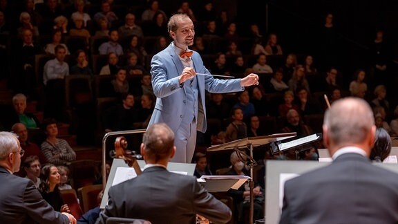 Alexander Schmitt dirigiert freudig im hellen Anzug beim Adventskonzert des MDR-Kinderchors auf der Bühne des Gewandhauses