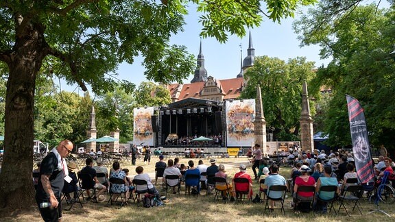 Das Publikum in sommerlicher Kleidung auf Klappstühlen schaut im Schlossgarten auf die Bühne, wo der MDR-Kinderchor vor der Kulisse des Merseburger Schlosses singt