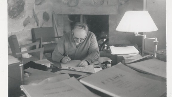 Thomas de Hartmann komponiert im Jahr 1950 über einen Tisch gebeugt, im Vordergrund verschiedene Partituren