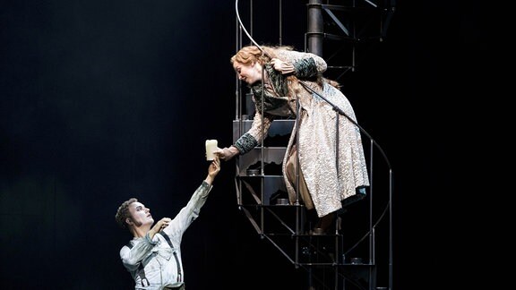 Ein Bühnenbild: Eine Frau in langem hellen Kleid steht auf einer Wendeltreppe aus Metall und beugt sich zu einem Mann herunter, der einen Arm nach ihrem ausstreckt