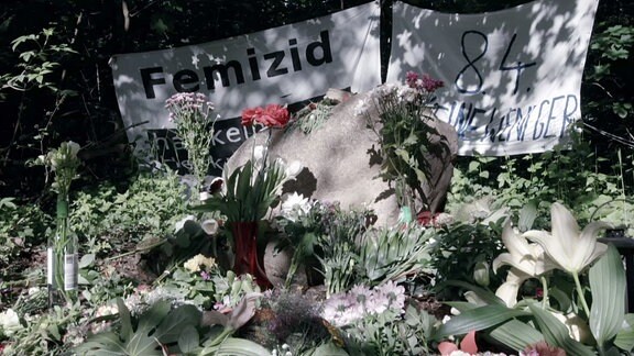 Blumen und Gedenkstein vor einem Plakat mit der Aufschrift Femizid.