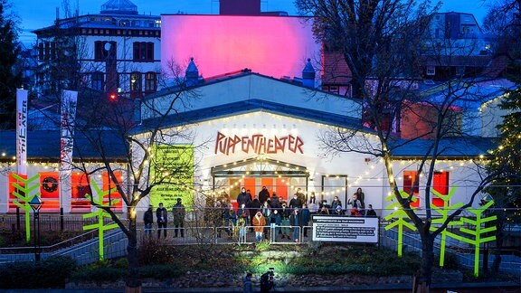 Mitarbeiter und Ensemble stehen vor dem mit Lichtern angestrahlten Puppentheater in Magdeburg.