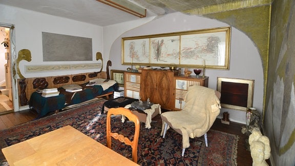 Ein Zimmer mit verschiedenen Wandmalereien, Bildern, einem bunten Sammelsurium an Möbeln und einem gemusterten Teppich