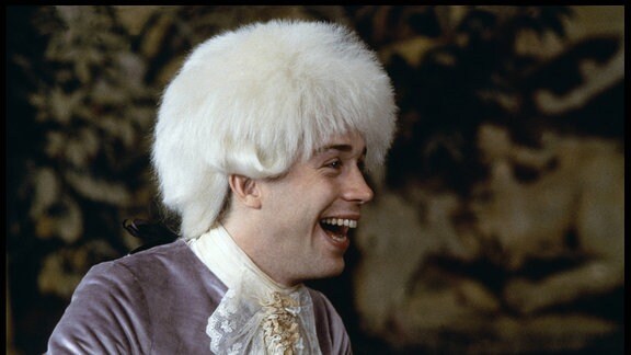 Ein jünger Mann mit einer weißen Perrücke lacht in einer Filmszene.