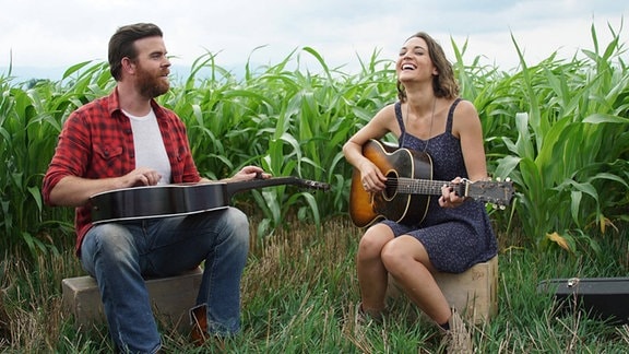 Zwei Personen sitzen mit Gitarren in einem Feld mit hochgewachsenen grünen Pflanzen.