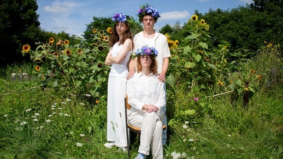 Auf einer grünen Wiese mit hohen Blumen sind drei Personen, eine sitzt, zwei stehen hinter ihr. Alle tragen weiße Kleidung.