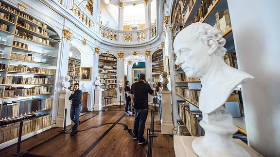 Zu sehen ist der Blick in den Rokokosaal der Herzogin Anna Amalia Bibliothek in Weimar. Der Raum ist in weiß und gold gehalten, Bücher stehen in den Regalen. Rechts im Bild ist eine weiße Büste zu sehen.