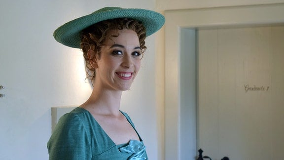 Eine Frau mit hochgesteckten Haaren und mit einem türkisen Sommerhut lächelt in die Kamera.