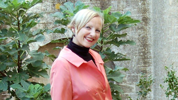 Eine Frau mit lachfarbener Bluse und schwarzem Rock steht an einer alten Betonmauer und lächelt in die Kamera.