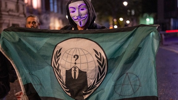Ein Mann mit Guy-Fawkes-Maske hält eine Fahne mit dem Anonymous-Logo hoch.