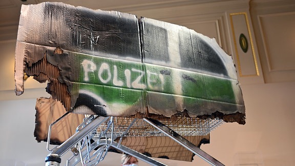 Einen Einkaufswagen, darauf befestigt eine Pappe mit darauf gemaltem Polizeiwagen.
