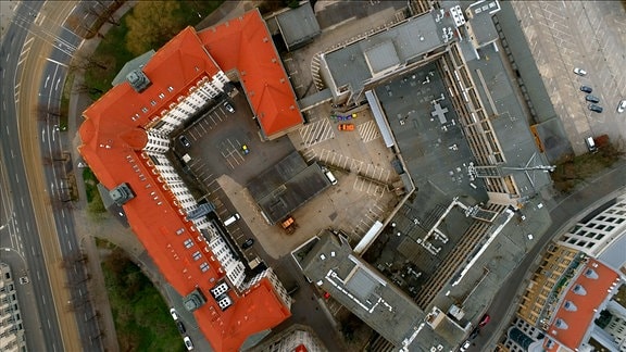 Blick von oben auf zwei Gebäude, eins mit rotem und eins mit grauen Dach, die jeweils in U-Form einen Platz umschließen. 
