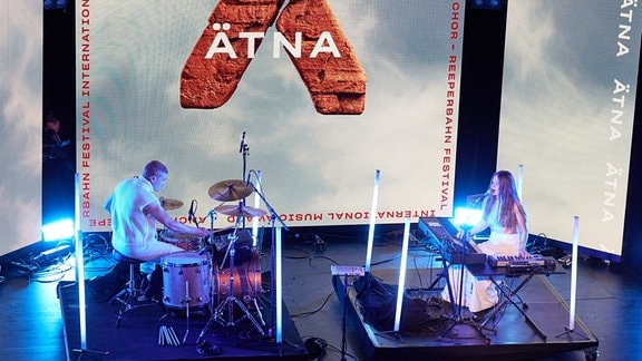 Ein Schlagzeuger sitzt auf einem Podest auf einer Bühne; ihm gegenüber sitzt eine Frau, die Keyboard spielt und singt