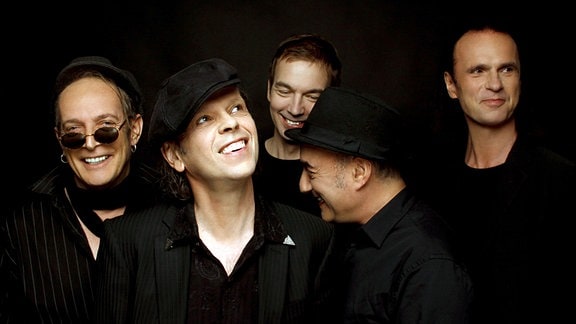 Fünf Männer der Band Pankow stehen in schwarzen Anzügen vor einem schwarzen Hintergrund.