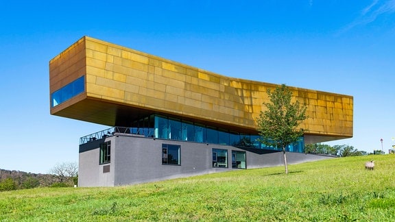 Blicke auf das Besucherzentrum der Himmelsscheibe Nebra: Gebäude auf einem grünen Hügel, obenauf eine goldener Gebäudeteil als eine Mischung aus einem rechteckigen Quader und einer Wiege.