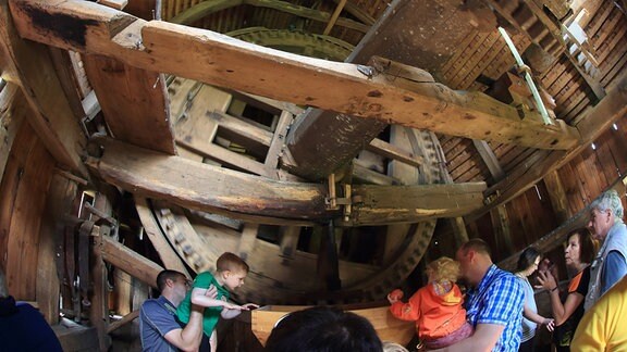 Blick in die hölzerne Maschinerie im Inneren einer Bockwindmühle; Erwachsene halten kleine Kinder auf dem Arm und helfen beim Hineinschauen
