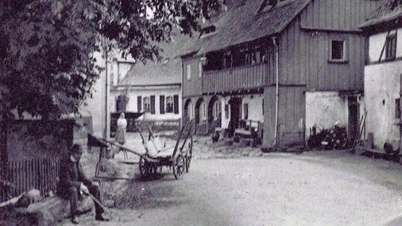 Historische Schwarz-Weiß-Aufnahme eines Bauernhauses mit angrenzender Scheune, davor ein leerer Heuwagen und ein Mann auf einer Bank unter einem Baum   