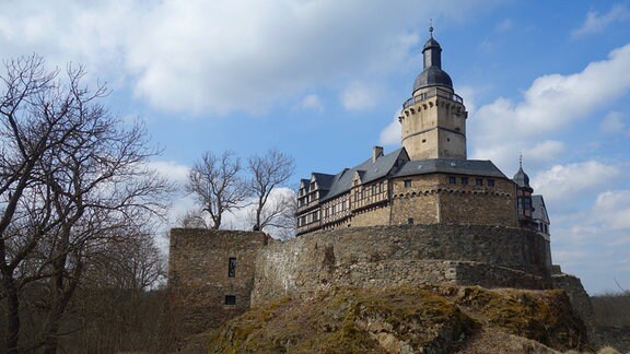 Zu sehen ist die Burg Falkenstein, leicht von unten fotografiert. Vorne sieht man Teile der Burgmauer, die Bäume daneben sind ohne Blätter