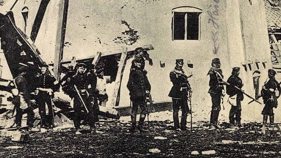 Eine alte Postkarte in schwarz-weiß zeigt preußische Soldaten vor der schwer beschädigten Düppler Mühle. Die Karte hat die Aufschrift "S.K.H. Prinz Friedrich Karl und Adjutanten an der Düppler Mühle", jemand hat handschriftliche Notizen hinzugefügt.
