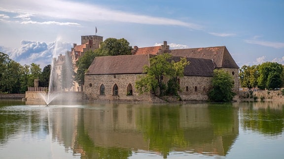 Hinter dem Schlossteich mit einer kleinen Wasserfontäne ist ein Gebäudeteil der Wasserburg Flechtingen zu sehen, er ist aus Natursteinen gebaut, im romanisch-frühgotischen Stil mit grau-roten Dachziegeln.