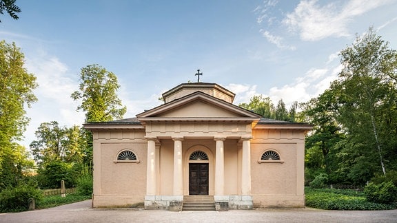 Zu sehen ist die Fürstengruft auf dem historischen Friedhof in Weimar. Das cremefarbene Mausoleum mit vier Säulen am Eingang und einem Kreuz auf dem Dach steht vor einigen grünen Bäumen, blauer Himmel