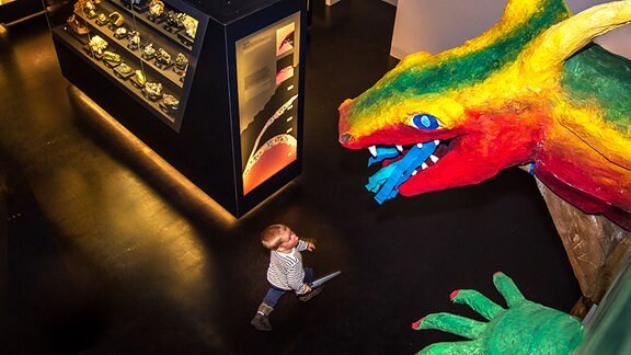 Kind mit Spielzeugschwert in der Hand blickt in einer Mineralienausstellung hinauf zu einer riesigen bunten Drachenfigur.