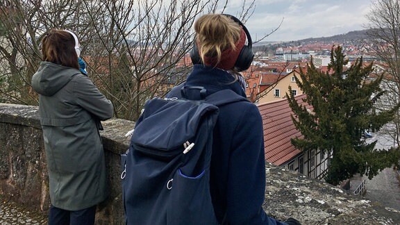 Zwei Frauen mit Kopfhöhrern von hinten an einem Aussichtspunkt mit Blick über eine Innenstadt.