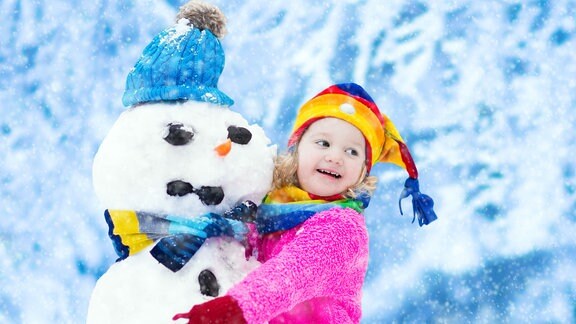 Fröhliches kleines Mädchen mit bunter Mütze und warmem Mantel, das mit einem Schneemann spielt. 