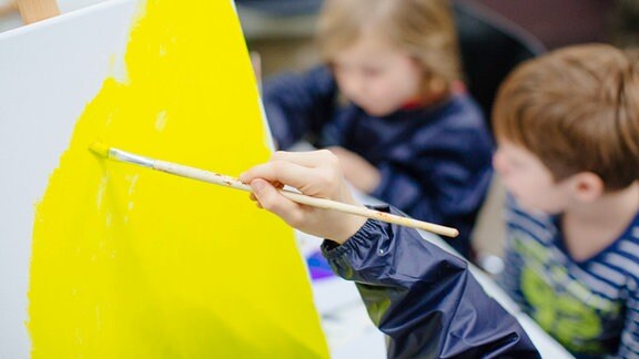 Eine Kinderhand malt mit einem Pinsel gelbe Farbe auf eine weiße Leinwand