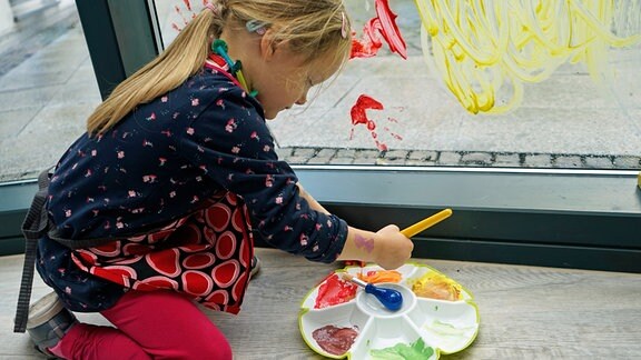 Ein blondes Mädchen sitzt vor einem mit Fingerfarben bemalten Fenster und taucht einen Pinsel in eine Farbpalette.