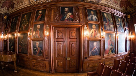 Aufnahme eines holzvertäfelten Bankettsaals in einem Schloss, in dem die Porträts historisch bedeutender Persönlichkeiten hängen.