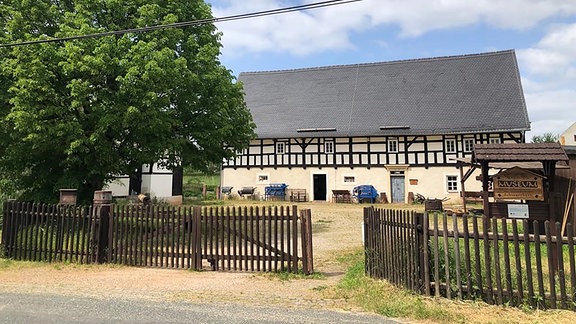 Ein altes Fachwerkhaus vor ihm liegt ein Hof der von einem Holzzaun begrenzt wird, links im Bild steht ein großer Baum, rechts sieht man die Beschilderung als Museum