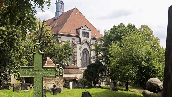 Ein Friedhof: im Vordergrund ein Kreuz mit verzierten Ecken, daneben ein Grabstein mit einem Engel. Im Hintergrund eine Kapelle.