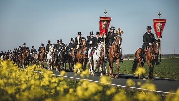 Zu sehen ist eine Prozession von Reitern, die Männer tragen schwarze Anzüge und Hüte, die vorderen Reiter tragen jeweils ein Banner