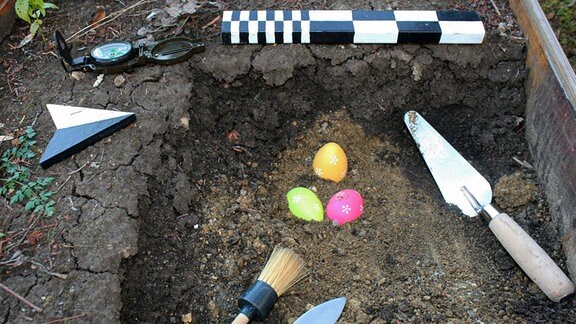 Zu sehen ist, wie Ostereier in einer Grabungsstätte liegen. Daneben liegt das entsprechende Werkzeug.