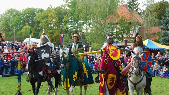 Vier Ritter sitzen in Rüstung auf ihren Pferden, sie tragen verschiedene farbenfrohe Wappen.