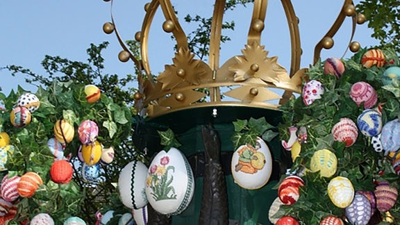 Zu sehen ist die Osterkrone an einem Brunnen in Greiz, an der Krone hängen verschieden gestaltete Ostereier.