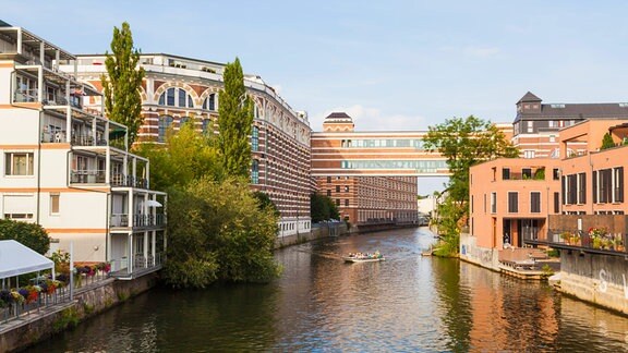 Der Fluss Weiße Elster in Leipzig, links und rechts stehen rot-weiße Gebäude. Im Hintergrund ein imposanter Fabrikbau. 