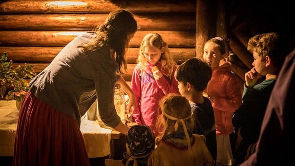 Kinder und eine Frau stehen in einer Blockhütte, trinken Tee und essen Kekse.