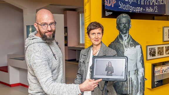 Ein Mann und eine Frau zeigen eine Mediaguide-App, die gehörlosen Menschen den Museumsbesuch ermöglicht.