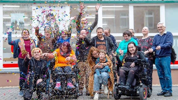 Eine Gruppe Menschen steht vor einem Gebäude, darunter auch mehrere Menschen im Rollstuhl, und werfen freudig mit Konfetti um sich.